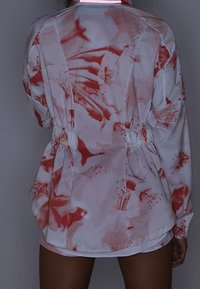Personne portant une veste blanche à manches longues avec des motifs abstraits rouges, resserrée à la taille, vue de dos.