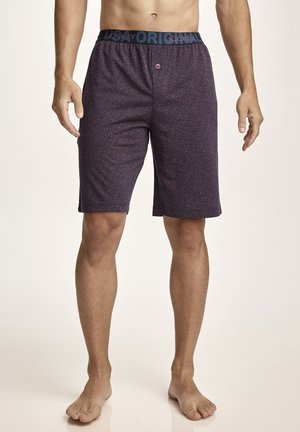 Mann trägt dunkel gemusterte knielange Shorts mit elastischem Bund und Markenzeichen, steht barfuß vor einem schlichten Hintergrund.