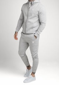 Grauer Reißverschluss-Hoodie mit Kängurutaschen und passenden Jogginghosen. Verfügt über eine Kapuze mit Kordelzug und ein Logo auf der Brust. Abgerundet mit weißen Sneakern.