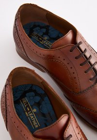 Chaussures habillées en cuir marron avec des détails brogue, un design à lacets et un talon texturé. L'intérieur présente une semelle intérieure à motif floral.