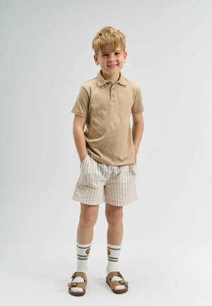 Short beige polo shirt, striped beige and white shorts, white socks with smiley face, and brown sandals. Standing confidently with hands in pockets.