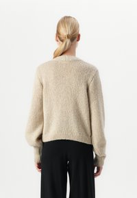 Blond kvinne med bakbundet hår iført en beige strikkegenser og svarte bukser, sett bakfra mot en ensfarget bakgrunn.