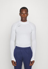 White long-sleeve athletic shirt with ribbed texture, featuring a high neckline and logo detail. Paired with navy pants with side stripes.