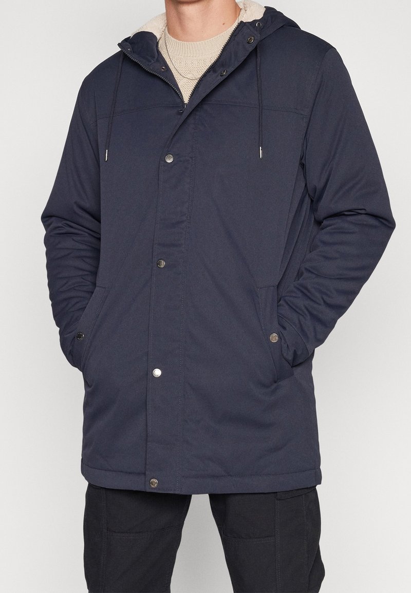 Mies, jolla on päällään tummansininen hupullinen talvitakki hopeisilla painikkeilla, alla beige neulepaita ja mustat housut, kädet taskuissa.