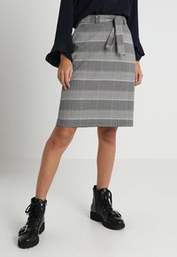 Grauer karierter Bleistiftrock mit Taillengürtel, gerade geschnitten und knielang. Kombiniert mit schwarzen Schnür-Stiefeletten.