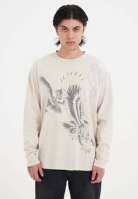Beige Langarm-T-Shirt mit schwarzem Adler-Graphic-Druck. Mit Rundhalsausschnitt, lockerer Passform und weichem Baumwollmaterial.