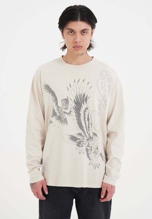 Beige Langarm-T-Shirt mit schwarzem Adler-Graphic-Druck. Mit Rundhalsausschnitt, lockerer Passform und weichem Baumwollmaterial.