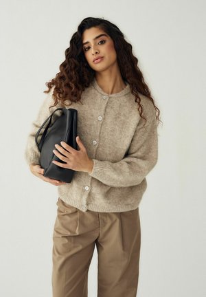 Kobieta o długich kręconych włosach, ubrana w beżowy sweter na guziki i brązowe spodnie, trzymająca czarną torebkę na jednolitym tle.