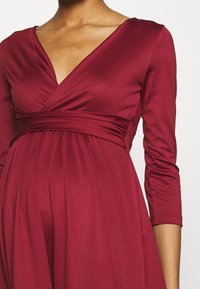 Vinröd mammaklänning med långa ärmar, V-ringning och rynkad midja. Mjukt tyg med en flödande silhuett.