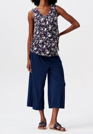 Kobieta ubrana w czarno-biały wzorzysty top bez rękawów, szerokie, granatowe spodnie o kroju cropped oraz czarne sandały wsuwane, stojąca z jedną ręką na biodrze.
