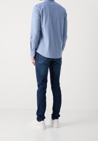 Uomo in piedi di spalle che indossa una camicia a maniche lunghe azzurro chiaro, jeans blu scuro e sneakers bianche su uno sfondo bianco uniforme.