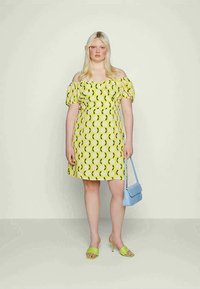 Vestido de ombros à mostra em tecido amarelo com padrão de limões verdes, mangas bufantes, cintura justa e saia rodada. Combinado com uma bolsa azul e saltos verdes.