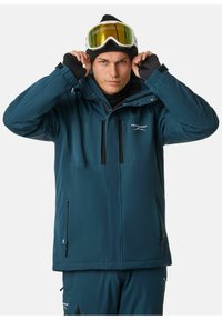 Tealfarbene Außenjacke mit Kapuze, mit Reißverschluss versehenen Taschen und schwarzen Akzenten; das Modell passt seine Skibrille an, während es eine schwarze Beanie und eine Basisschicht trägt.