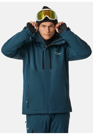 Tealfarbene Außenjacke mit Kapuze, mit Reißverschluss versehenen Taschen und schwarzen Akzenten; das Modell passt seine Skibrille an, während es eine schwarze Beanie und eine Basisschicht trägt.