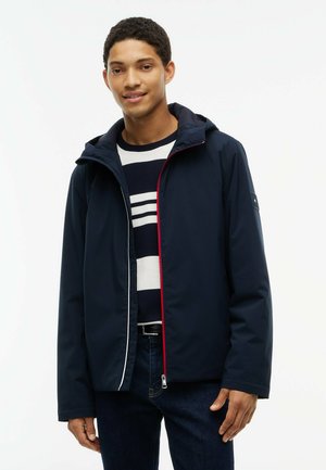 Ung mand iført en navyjakke med hætte over en sort og hvid stribet sweater og mørke jeans, stående foran en ensfarvet hvid baggrund.