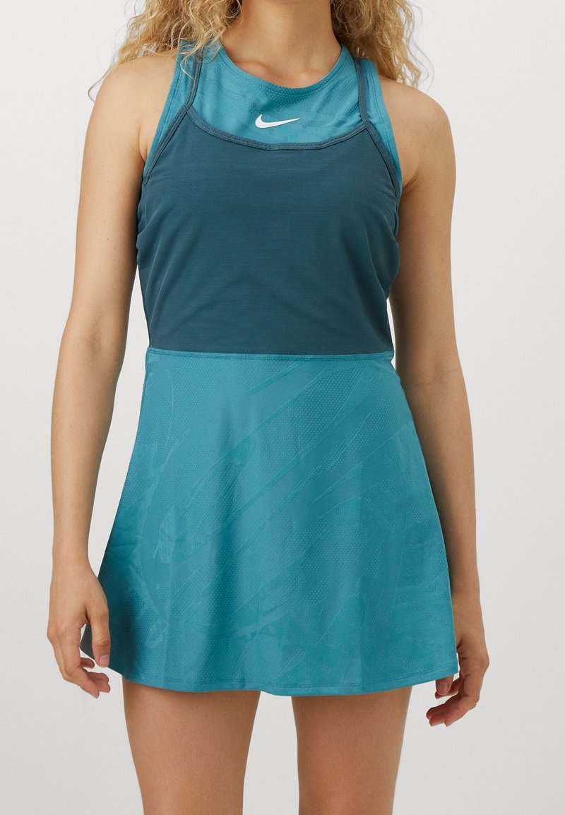 Femme portant une robe de sport sans manches de couleur sarcelle avec le logo Nike sur la poitrine, debout devant un fond clair uni.