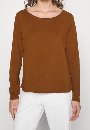 Vrouw draagt een bruin shirt met lange mouwen en een ronde hals, en een witte broek, staand tegen een effen lichte achtergrond.