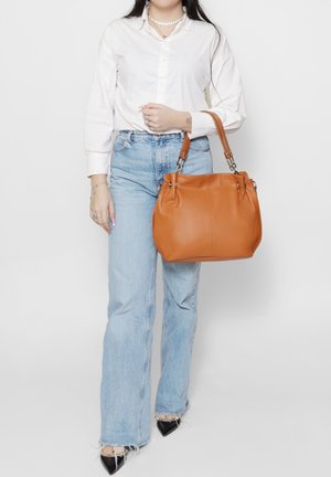 Borsa in pelle color cuoio con finitura liscia, forma arrotondata, doppi manici e dettagli in metallo. Abbinata a una camicia bianca e jeans azzurri.