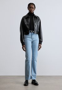 Prerezana crna kožna jakna sa zatvaračem, uparena sa svijetloplavim trapez hlačama. Model nosi crni rolka i crne cipele.