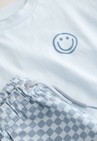 Světle modré tričko s modrým smajlíkem, doplněné modro-bílými kostkovanými šortkami se šňůrkou.