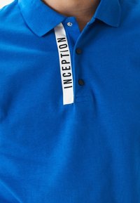 Modrá polo košile s texturovanou tkaninou, s bílou etiketou s nápisem "INCEPTION" na légě a dvěma černými knoflíky.