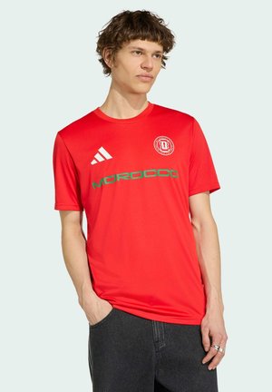 Ung mand iført en rød Marokko fodboldtrøje med Adidas-logo og grøn skrift, stående med den ene hånd i lommen.