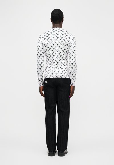 Man standing facing away wearing a white long-sleeve shirt with black crescent patterns and black loose pants with black shoes.