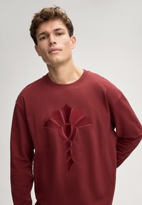 Felpa bordeaux con colletto rotondo, ricamo testurizzato di un design stilizzato al centro, realizzata in morbido tessuto, maniche lunghe.