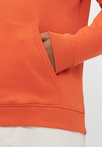 Mano nella tasca anteriore di una felpa arancione con polsini e orlo a coste, sopra pantaloni di colore chiaro.