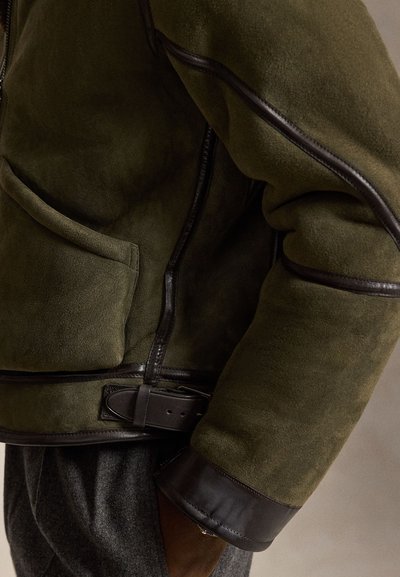 Grüne Wildlederjacke mit dunklem Lederbesatz, Gürtel-Detail, Manschettenakzenten und seitlicher Tasche, getragen über grauen Wollhosen.