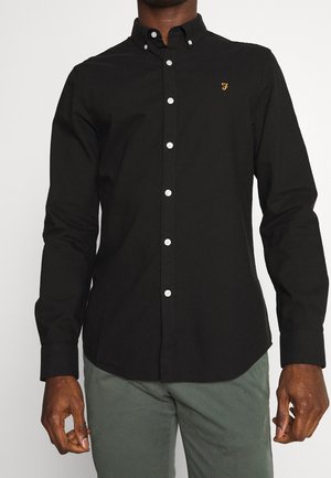 Hombre con una camisa negra ajustada abotonada con un pequeño logo dorado en el pecho y pantalones verdes, de pie con los brazos relajados a los lados.