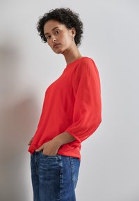 Rote Bluse mit langen, gerafften Ärmeln, kombiniert mit blauen Jeans. Der Stoff hat eine strukturierte Optik, die Brusttaschen sind nicht sichtbar.
