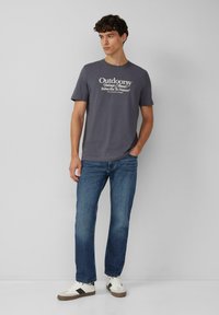 Grijze katoenen t-shirt met korte mouwen en een ronde halslijn, voorzien van een witte tekst grafisch ontwerp. Gecombineerd met blauwe jeans en witte sneakers.