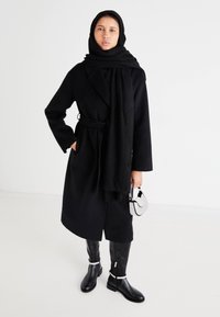 Manteau noir avec ceinture, porté sur une tenue noire. Porte une longue écharpe noire. Tient un petit sac à motif. Bottes noires montantes jusqu'aux genoux.