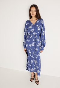 Μπλε φλοράλ φόρεμα με μακριά μανίκια και βαθύ V-λεκέ. Διαθέτει εφαρμοστόed μέση και ροή σ' ανοιχτό φόρεμα. Μαύρες σαγιονάρες με λουράκια και μαύρη τσάντα clutch.