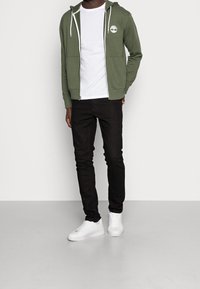 Sudadera con cremallera verde oliva y cordones blancos, con un logo blanco en el pecho, llevada sobre una camiseta blanca, combinada con jeans negros y zapatillas blancas.
