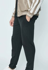 Veste bomber pour homme en beige avec des rayures blanches, associée à un pantalon noir. La veste dispose de poignets côtelés et de boutons-pression. Baskets blanches.