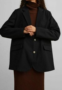 Schwarze Blazer mit maßgeschneidertem Design, zwei vorderen Taschen und hellen Knöpfen. Getragen über einem gerippten braunen Rollkragenkleid.