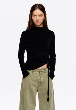 Top de terciopelo negro de manga larga con cuello alto, frunce lateral y detalle de lazo; combinado con pantalones verdes olivo de corte holgado.