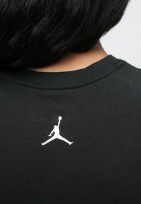 Άτομο με σκούρα μαλλιά που φοράει ένα μαύρο πουκάμισο με λευκή σιλουέτα ενός παίκτη μπάσκετ που πηδάει με μια μπάλα κοντά στον λαιμό.