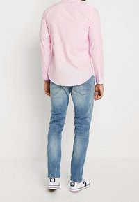 Camicia rosa a maniche lunghe con colletto, abbinata a jeans azzurri. Le scarpe sono bianche con un logo blu navy, con un design classico da sneaker.
