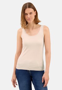 Donna con capelli castano chiaro che indossa un top beige senza maniche e jeans blu, in piedi contro uno sfondo leggero e semplice.