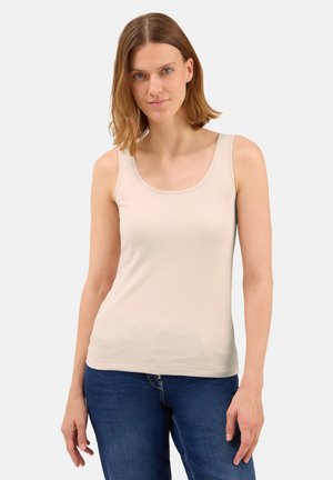 Femme aux cheveux châtain clair portant un haut sans manches beige et un jean bleu, se tenant devant un fond lumineux uni.