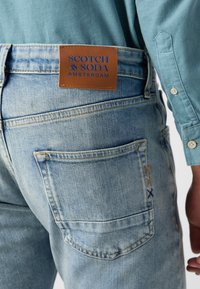 Jeans de mezclilla azul claro con una etiqueta de cuero marrón que dice "SCOTCH & SODA AMSTERDAM" en la cintura y costuras decorativas en los bolsillos.