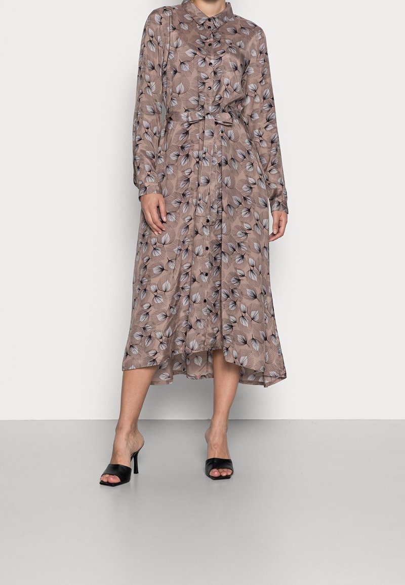 Robe marron à manches longues avec un motif de feuilles beige et noir, boutonnée devant, ceinture à la taille, et chaussures noires à talons hauts ouvertes.