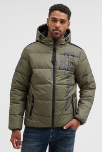 Uomo che indossa una giacca imbottita verde oliva con cappuccio, cerniera blu navy e testo stampato, in piedi con una mano in tasca davanti a uno sfondo semplice.