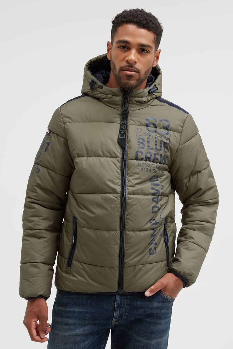 Uomo che indossa una giacca imbottita verde oliva con cappuccio, cerniera blu navy e testo stampato, in piedi con una mano in tasca davanti a uno sfondo semplice.