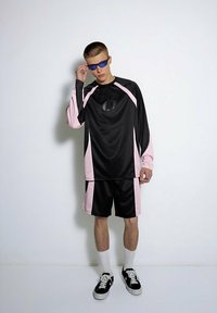 Czarny i różowy strój sportowy składa się z koszulki z długim rękawem o prążkowanej strukturze z logo, w komplecie z pasującymi szortami. Zestaw zawiera również okulary przeciwsłoneczne.