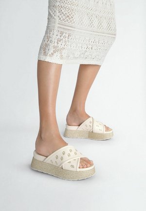 Sandales à plateforme beiges avec des brides croisées et des accents décoratifs dorés sur la partie supérieure. Semelle texturée et scintillante ajoutant de la dimension.