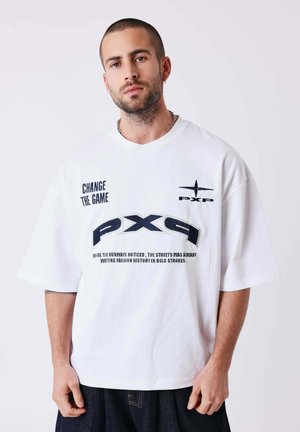 Homme aux cheveux courts et barbe porte une chemise blanche oversize avec du texte noir "CHANGE THE GAME" et des logos "PXP" sur un fond uni.